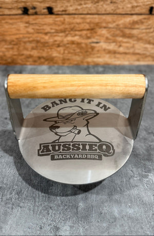 Metal protein press with wooden handle on a gray surface, featuring 'AussieQ Backyard BBQ' branding.
